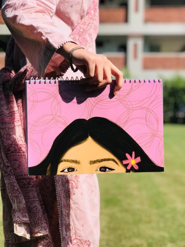 A landscape-oriented pink spiral notebook featuring a hand-painted illustration of the top half of a girl's face with deep brown eyes and a small pink flower in her dark hair.