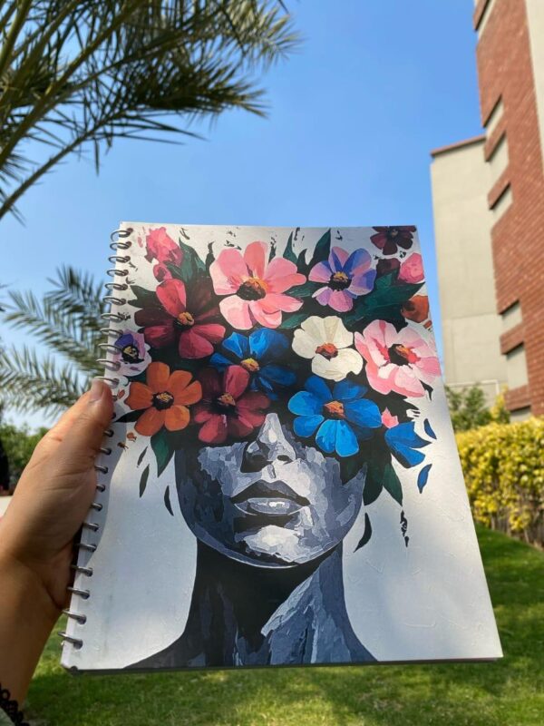 A spiral-bound notebook featuring a bold, colorful painting of a person’s face with a large, vibrant crown of blooming flowers.