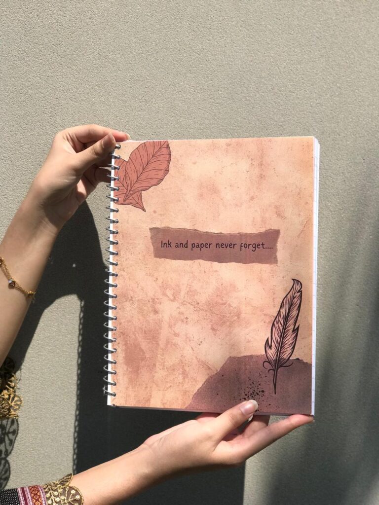 A vintage-themed spiral notebook with a rustic brown textured cover, featuring two feather illustrations and the quote "Ink and paper never forget...." in the center.