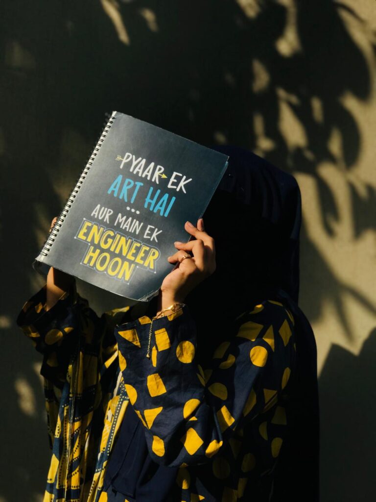 A dark-themed spiral notebook held by a person, featuring the funny text "PYAAR EK ART HAI... AUR MAIN EK ENGINEER HOON"